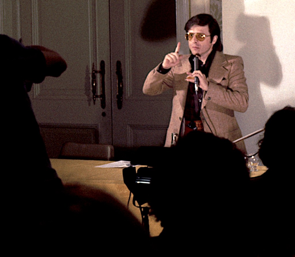 Harlan Ellison speaks at a panel discussion, framed by members of the audience. 