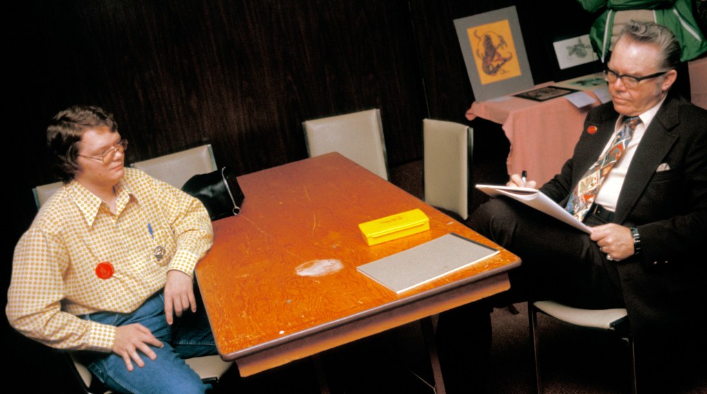 A medium shot of artist Kelly Freas sitting at a folding table, sketchbook on his knee, drawing a young man opposite. 