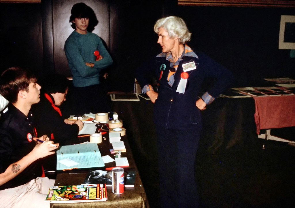 Elizabeth Pearse talking to convention volunteers, probably in the art-show room although the location is unclear. 