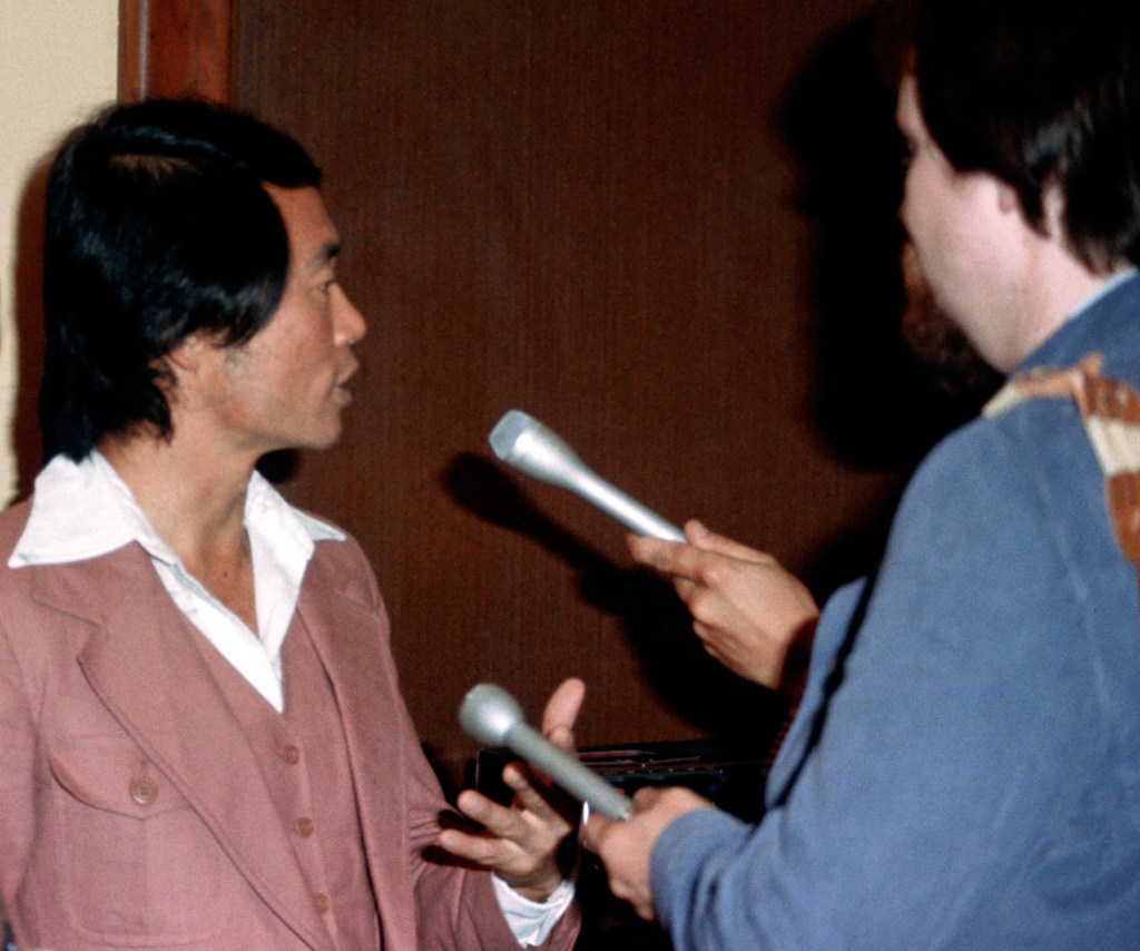 George Takei, wearing a very 70s suit, is seen being interviewed by two people holding microphones. 